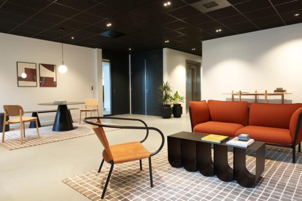 Moderne wachtruimte met designmeubilair, waaronder een oranje bank, zwarte salontafel en houten stoelen op een geruit tapijt.